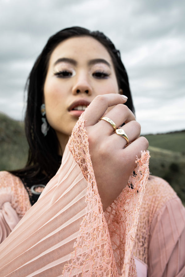 model wears peach coloured dress with pleated sleeves, and two gold rings. She is holding her hand up to the camera